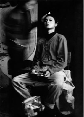 a black and white photograph of what appears to be a young Chinese man (the artist Ma Liuming) wearing trousers and a buttoned up short and sitting with legs spread on a chair. Next to him is the blurred figure of another person in movement.
