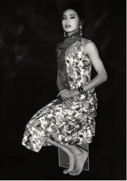 A black and white photograph of what appears to be a young Chinese female (the artist Ma Liuming) wearing an elegant white dress with a flower pattern and a scarf draped around his neck and side body. His perches on a chair with knees closed together and the hands clasped. He also wears a bangle and very long ear rings. His mouth is slightly open.