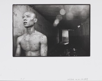 A black and white photograph showing the naked torso and shaved head of a young Chinese man (the artist Zhang Huan) covered in sweat and flies. Behind him there is a dark concrete room with a rectangular doorway at the end. The floor looks wet.